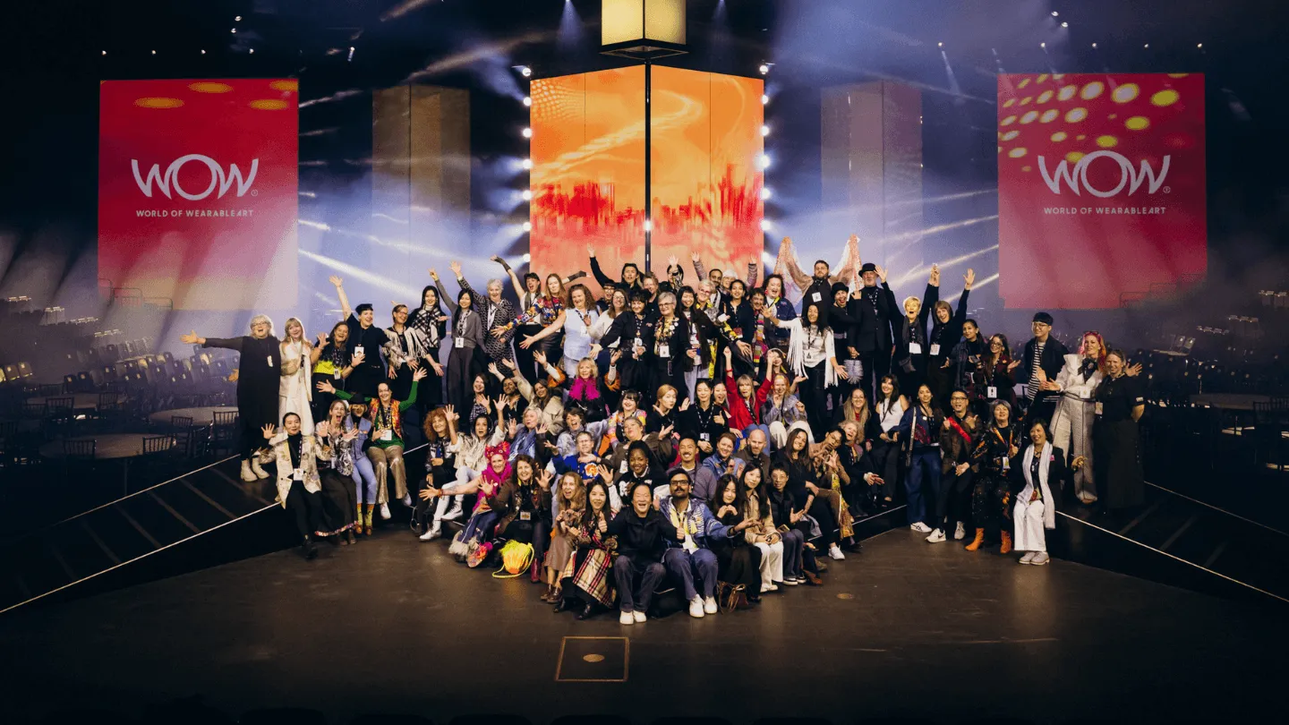Large group of diverse people posing together on stage with raised hands under bright lights and WOW World of Wearable Art banners.